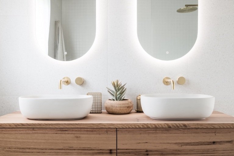 Moderne badeværelser med vaskeskab i træ og håndvask og badeværelsesbeslag i guldoptik og spejle med belysning og hvide granitfliser